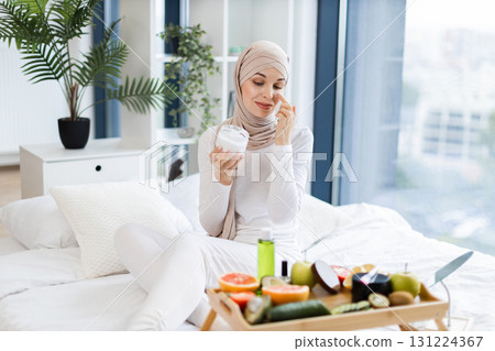 Smiling young Muslim woman wearing hijab applying moisturizing cream to face, sitting on bed in bright modern rooms, surrounded by skincare and fresh fruits showcasing self-care, skincare 131224367
