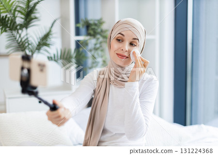 Young Muslim woman in light-colored hijab using cotton pads, blogging about skincare routine and personal health, sitting in an elegant and sunlit room while capturing content on a mobile device. 131224368