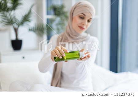 Young Muslim woman wearing hijab applying lotion for skincare in bright modern room. Focusing on self-care beauty and healthy lifestyle. Vibrant natural light and greenery create peaceful atmosphere. 131224375