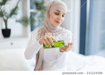 Young Muslim woman wearing beige hijab cleansing her face with lotion and cotton pad. Seated in modern bright home environment with plants in background, practicing skincare routine Young Muslim woman wearing beige hijab cleansing her face with lotion and cotton pad. Seated in modern bright home environment with plants in background, practicing skincare routine 131224380