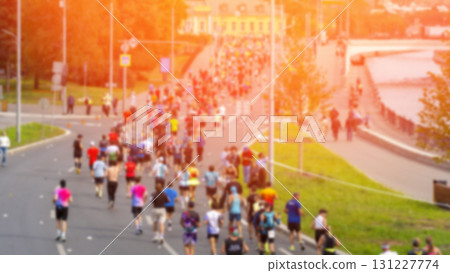 The throng of runners moves in a blur as they traverse the verdant city streets, the golden light casting a warm and vibrant glow. The crowd of runners moves in unison during the marathon along the The throng of runners moves in a blur as they traverse the verdant city streets, the golden light casting a warm and vibrant glow. The crowd of runners moves in unison during the marathon along the 131227774