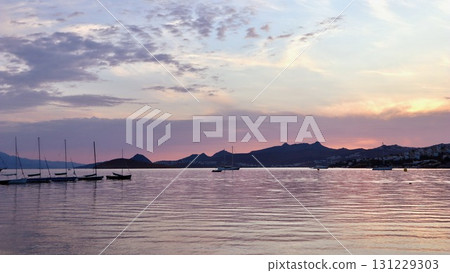 A peaceful coastal sunset with soft pink and purple clouds reflected on the calm water, with a silhouette of distant hills and a line of sailboats. 131229303