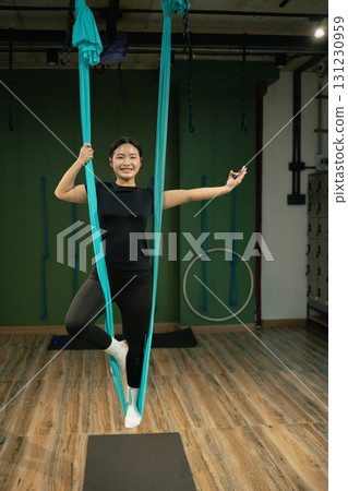 Smiling woman practicing aerial yoga, standing in hammock support while posing with arms open 131230959