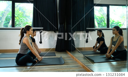 Two women practicing seated butterfly pose on yoga mats in a bright studio with mirror reflection Two women practicing seated butterfly pose on yoga mats in a bright studio with mirror reflection 131231084