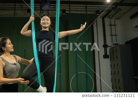 Yoga instructor assisting woman practicing aerial yoga with hammock support in fitness studio 131231121