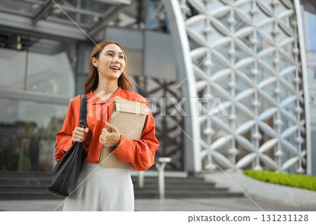 Confident young businesswoman standing outside office building holding documents and smiling 131231128