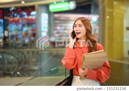 Businesswoman talking on mobile phone while holding documents inside modern shopping mall 131231130