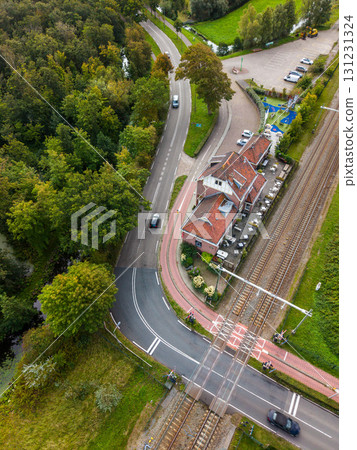 Aerial view of a diagonal railway crossing in a semi rural area, with cars halted at barriers, a red roofed building with outdoor seating, green fields, trees, and a nearby parking lot. Aerial view of a diagonal railway crossing in a semi rural area, with cars halted at barriers, a red roofed building with outdoor seating, green fields, trees, and a nearby parking lot. 131231324