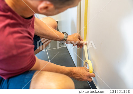 Man applying masking tape to baseboards while preparing walls for white paint. Home renovation, precision work and interior improvement during DIY construction Man applying masking tape to baseboards while preparing walls for white paint. Home renovation, precision work and interior improvement during DIY construction 131231338