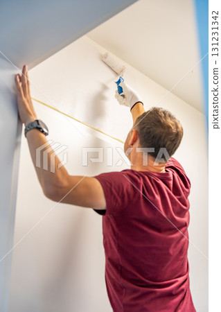 Man painting the wall with a roller in white during home renovation. Interior improvement, surface transformation and DIY work with paint tools. 131231342