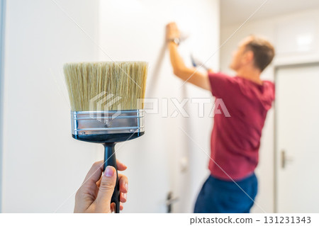 Close-up of a hand holding a paintbrush with a man painting the wall in the background. Concept of manual work, home renovation and attention to detail during interior improvement. Close-up of a hand holding a paintbrush with a man painting the wall in the background. Concept of manual work, home renovation and attention to detail during interior improvement. 131231343
