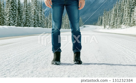 A person wearing jeans and winter boots stands on a snow-covered road, surrounded by a snowy winter landscape 131232384