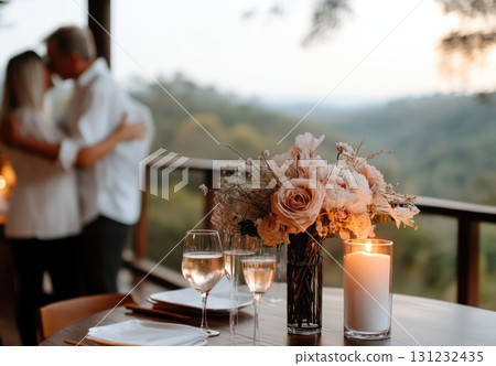 Romantic couple embraces in the background, while a candlelit table set with wine glasses and flowers creates an intimate ambiance on a balcony overlooking scenic mountains Romantic couple embraces in the background, while a candlelit table set with wine glasses and flowers creates an intimate ambiance on a balcony overlooking scenic mountains 131232435