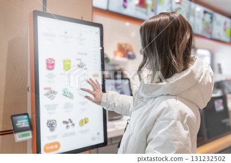 A 30 - 35-year-old woman in a white winter jacket orders cakes and food through a touchscreen screen in a fast food restaurant or pizzeria coffee shop. Person Using Interactive Kiosk for Ordering in a 131232502