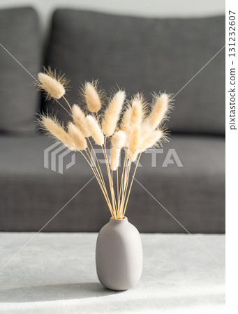 Dried grass "rabbit tail" in a matte vase standing on a concrete table against a soft gray sofa. Dried grass "rabbit tail" in a matte vase standing on a concrete table against a soft gray sofa. 131232607