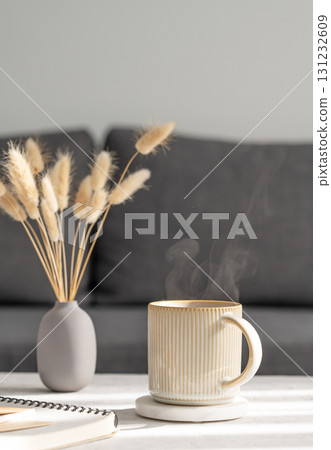 Beige ceramic mug with steam on a light table with soft shadows, vase and notebook on the background Beige ceramic mug with steam on a light table with soft shadows, vase and notebook on the background 131232609