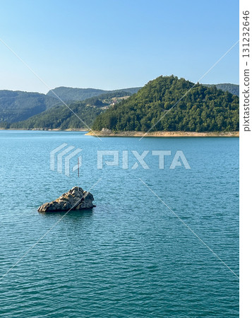 Rocky islet with a cross in peaceful blue lake, surrounded by green forest hills under clear sky 131232646
