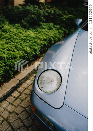 Vintage blue car headlight gleaming beside green foliage, resting on cobblestone pavement, highlighting automotive styling and nostalgic design details. Close up view of headlight of vintage car Vintage blue car headlight gleaming beside green foliage, resting on cobblestone pavement, highlighting automotive styling and nostalgic design details. Close up view of headlight of vintage car 131232966