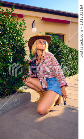 Stylish woman in cowboy hat kneeling with scissors and pliers near green bushes 131233164