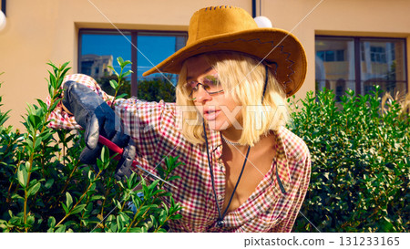 Stylish woman in cowboy hat wearing gloves trimming green bush with focused expression 131233165