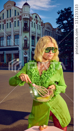 Blonde woman in green dress mixing eggs in glass bowl with stylish attitude outdoors 131233183