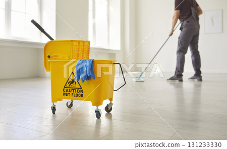 Yellow mop bucket with Caution Wet Floor sign with blue cloth and mop, man mopping empty room Yellow mop bucket with Caution Wet Floor sign with blue cloth and mop, man mopping empty room 131233330