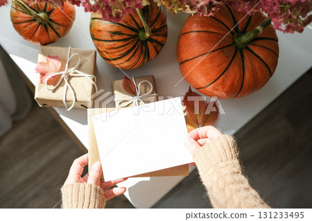 Cozy autumn flat lay with hands in sweater holding blank greeting card, surrounded by pumpkins, wrapped gifts and fall leaves on white surface. Ideal for seasonal greetings and Thanksgiving Cozy autumn flat lay with hands in sweater holding blank greeting card, surrounded by pumpkins, wrapped gifts and fall leaves on white surface. Ideal for seasonal greetings and Thanksgiving 131233495