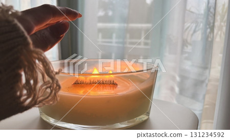 Large scented candle in a glass jar, a woman's hand warming up, cozy autumn home comfort and warm hygge lifestyle, aromatherapy Large scented candle in a glass jar, a woman's hand warming up, cozy autumn home comfort and warm hygge lifestyle, aromatherapy 131234592