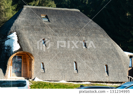 Exploring a charming thatched cottage nestled in a serene landscape during a clear winter day surrounded by snow and evergreen trees 131234995