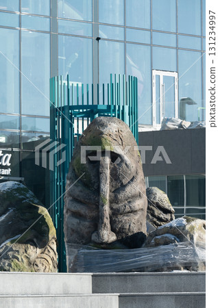 A large stone sculpture resembling a mythical creature stands prominently in an urban plaza surrounded by modern architecture under bright sunlight in the early afternoon 131234997
