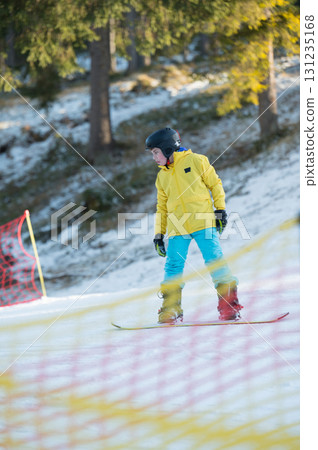 Brightly clad youth enjoys skiing adventure on a sunny winter day at a sloping terrain surrounded by snow-covered trees and colorful netting for safety 131235168