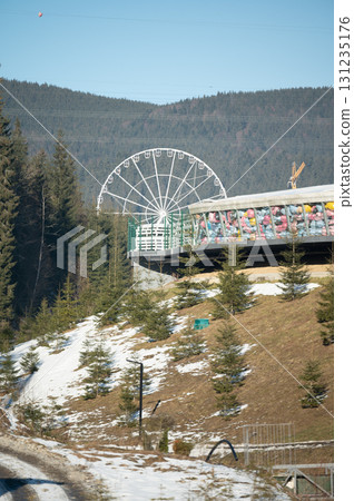 Winter landscape showcasing a colorful ferris wheel nestled among evergreen trees near a hilltop during a bright, clear day 131235176