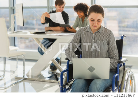Accessible environment. Woman in chair for people with disability working with laptop in modern office. Diverse multiracial group of people colleagues working together. Young professional team 131235667