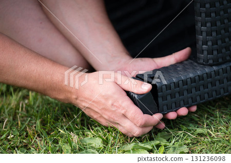 Assembling outdoor furniture on the grass. Person's hands carefully connect black woven pieces. Assembling outdoor furniture on the grass. Person's hands carefully connect black woven pieces. 131236098