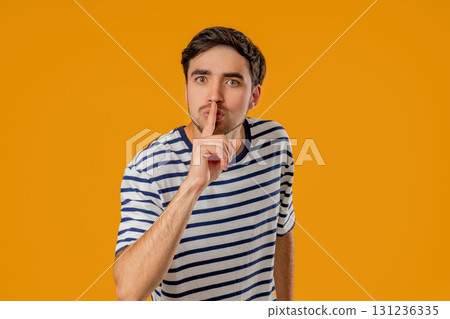 Handsome man with gesture of shhh, secret, silence, conspiracy, quiet, rumor concept. Smiling guy holding finger on lips, yellow studio background 131236335