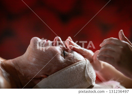 Close up of a mature woman lying on a treatment bed with a towel and headband while a professional therapist applies foam during a facial cleansing procedure. Skincare and wellness concept Close up of a mature woman lying on a treatment bed with a towel and headband while a professional therapist applies foam during a facial cleansing procedure. Skincare and wellness concept 131236497