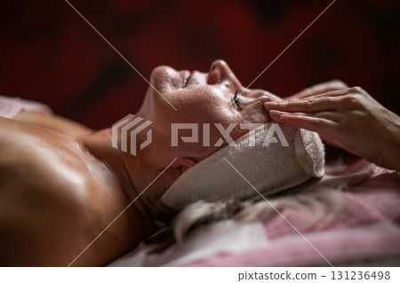 Side view close up of a mature woman lying on a treatment bed with a headband while a professional therapist massages her face. 131236498