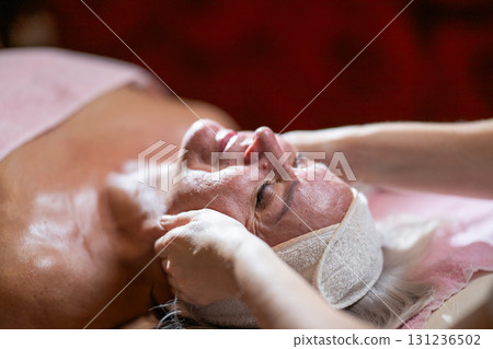Close up of a mature woman lying on a treatment bed with a headband while a therapist gently massages her face. Spa beauty and wellness concept focusing on relaxation, skincare, rejuvenation, and Close up of a mature woman lying on a treatment bed with a headband while a therapist gently massages her face. Spa beauty and wellness concept focusing on relaxation, skincare, rejuvenation, and 131236502