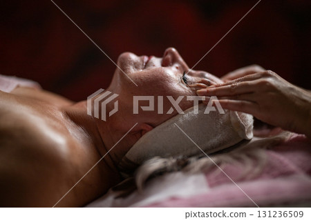 Side view close up of a mature woman lying on a treatment bed with a headband while a professional therapist massages her face. Side view close up of a mature woman lying on a treatment bed with a headband while a professional therapist massages her face. 131236509