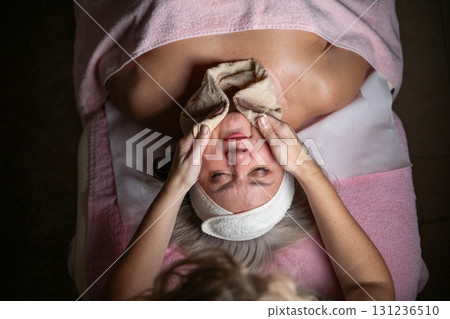 Close up of a mature woman lying on a treatment bed with a towel and headband while a professional therapist applies foam during a facial cleansing procedure. Skincare and wellness concept 131236510