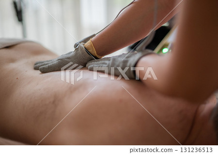 Close up view of a therapist applying electrotherapy with special conductive gloves on a man back during a physiotherapy session. Concept of modern rehabilitation, pain relief, muscle stimulation, and Close up view of a therapist applying electrotherapy with special conductive gloves on a man back during a physiotherapy session. Concept of modern rehabilitation, pain relief, muscle stimulation, and 131236513