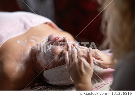 Close up of a mature woman lying on a treatment bed with a towel and headband while a professional therapist applies foam during a facial cleansing procedure. Skincare and wellness concept 131236519