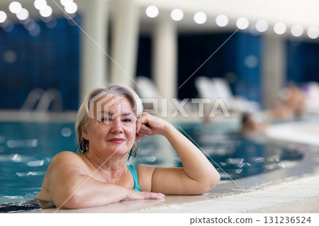 Smiling senior woman enjoying relaxation in an indoor swimming pool, leaning on the poolside. Concept of active lifestyle, wellness, health, hydrotherapy, and leisure for older adults. 131236524
