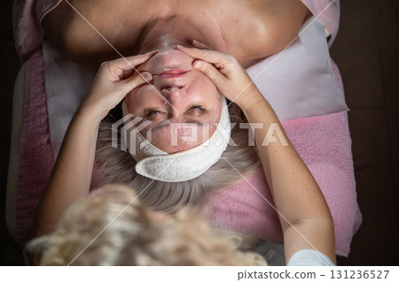 Close up of a mature woman receiving a relaxing facial massage from a professional female therapist in a modern spa. The client lies on a treatment bed with a towel and headband, enjoying skincare 131236527