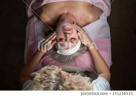 Close up of a mature woman lying on a treatment bed with a towel and headband while a professional therapist applies foam during a facial cleansing procedure. Skincare and wellness concept 131236533
