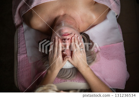 Close up of a mature woman receiving a relaxing facial massage from a professional female therapist in a modern spa. The client lies on a treatment bed with a towel and headband, enjoying skincare 131236534