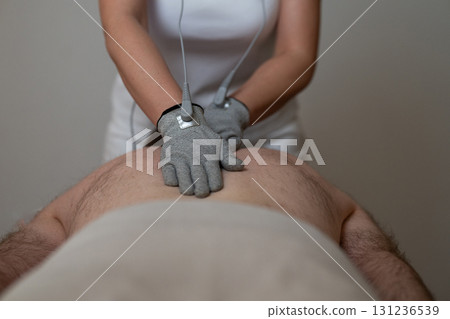 Close up view of a therapist applying electrotherapy with special conductive gloves on a man back during a physiotherapy session. Concept of modern rehabilitation, pain relief, muscle stimulation, and Close up view of a therapist applying electrotherapy with special conductive gloves on a man back during a physiotherapy session. Concept of modern rehabilitation, pain relief, muscle stimulation, and 131236539