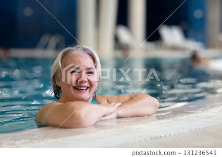 Smiling senior woman enjoying relaxation in an indoor swimming pool, leaning on the poolside. Concept of active lifestyle, wellness, health, hydrotherapy, and leisure for older adults. Smiling senior woman enjoying relaxation in an indoor swimming pool, leaning on the poolside. Concept of active lifestyle, wellness, health, hydrotherapy, and leisure for older adults. 131236541