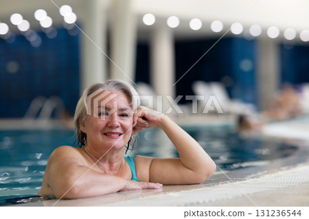 Smiling senior woman enjoying relaxation in an indoor swimming pool, leaning on the poolside. Concept of active lifestyle, wellness, health, hydrotherapy, and leisure for older adults. 131236544