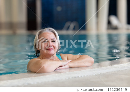 Smiling senior woman enjoying relaxation in an indoor swimming pool, leaning on the poolside. Concept of active lifestyle, wellness, health, hydrotherapy, and leisure for older adults. Smiling senior woman enjoying relaxation in an indoor swimming pool, leaning on the poolside. Concept of active lifestyle, wellness, health, hydrotherapy, and leisure for older adults. 131236549